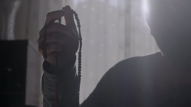 Backlit by soft sunlight, a man gently holds rosary beads during a moment of meditation. Themes of inner peace, spiritual grounding, resilience, and contemplation.