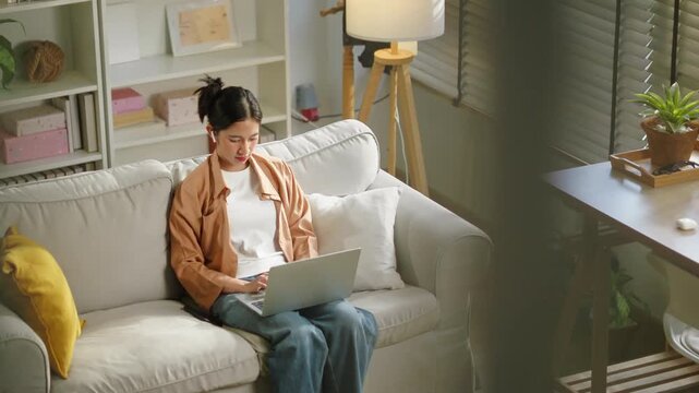 Happy Asian woman using laptop for video meeting on sofa at home, remote working corporate employee, digital nomad hybrid workplace business executive and flexible lifestyle technology concept