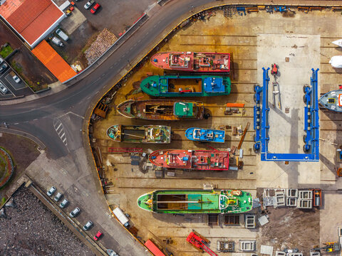Aerial view of colorful fishing boats undergoing maintenance in a dry dock shipyard with a blue boat lift and paved surroundings in Madalena, Azores, Portugal.