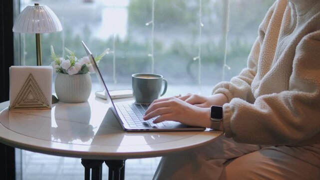Person types on laptop at cozy table near window. Warm light illuminates workspace with coffee and small plants. Scene suggests calm focus or remote work. Ideal for business, education