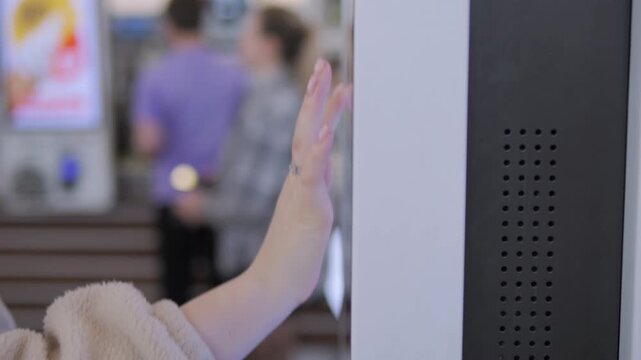 Hand taps interactive kiosk screen. Background shows blurred people. Scene suggests modern retail or public space. Useful for tech interfaces or customer service clips. No visible emotions on people