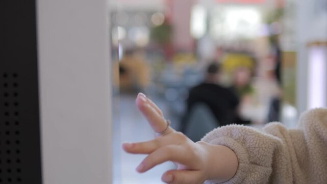 Child's hand reaches toward interactive screen in modern cafe. Focus on tiny fingers, soft sweater, blurred background. Quiet curiosity, gentle movement, playful exploration