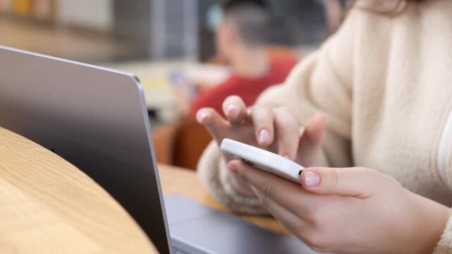 Close-up of a person's hands using a smartphone. Laptop sits open beside them on wooden table. Blurred background shows another person seated nearby. Focus remains on fingers scrolling