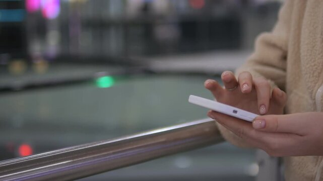 Person holds phone while standing on moving walkway. Focus on hands and device, background blurred. Neutral expression, casual travel moment. Suitable for travel, technology, or urban transit themes