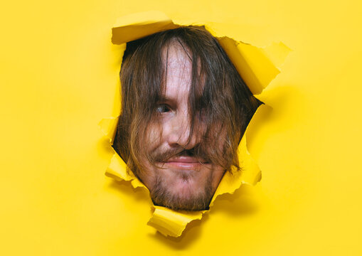 A long-haired, shaggy man with a beard and mustache looks out from a torn hole in yellow paper with a strange expression on his face and a sly smile. The concept originality,old-fashioned, originality
