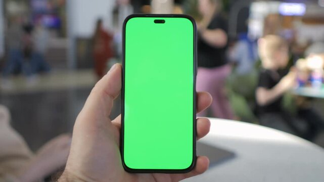 Hand holds phone with green screen. Background shows blurred cafe scene. Person appears focused, calm. Footage useful for app previews or mockups. Great for digital marketing projects