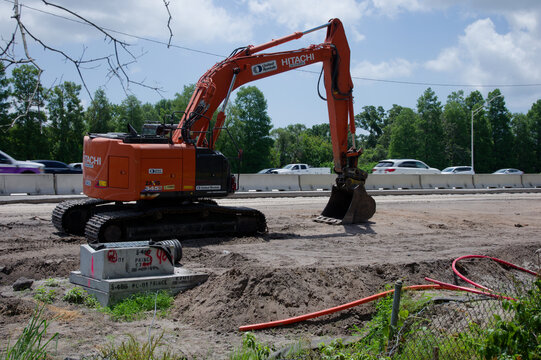 Orange Hitachi excavator working on a roadside construction site, digging and leveling earth. Editorial Use Only June 24, 2026 St. Petersburg, FL USA beside concrete barriers with passing highway traf