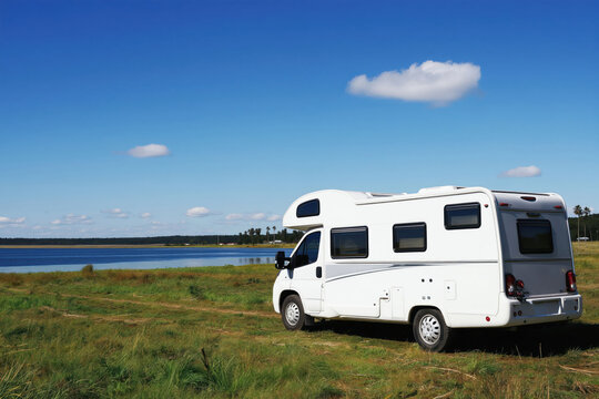 White motorhome parked on grassy field next to calm lake under blue sky. Leisure vehicle awaits campers for outdoor adventures, family trips, and nature holidays.
