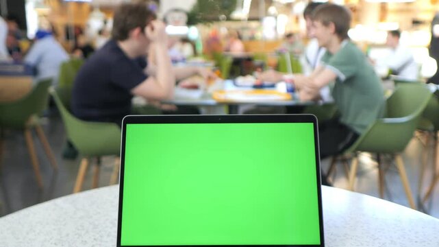Laptop with green screen sits on cafe table. Background shows blurred people chatting and eating. Focus remains on laptop's screen. Useful for digital content, presentations, or mockups