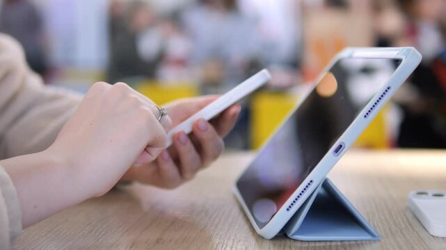 Close-up of hands using tablet and smartphone on wooden table. Soft focus background shows casual indoor setting. Person appears focused, engaged with digital devices. Ideal for tech, education