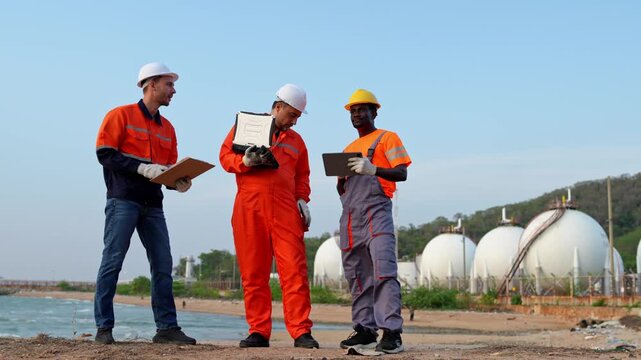 Team of industrial workers examining blueprints at an oil storage site with a clear blue sky overhead