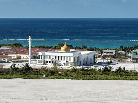 Aerial view of a white mosque with a golden dome surrounded by lush tropical vegetation and land reclamation in Dhidhdhoo, Haa Alif Atoll, Maldives.