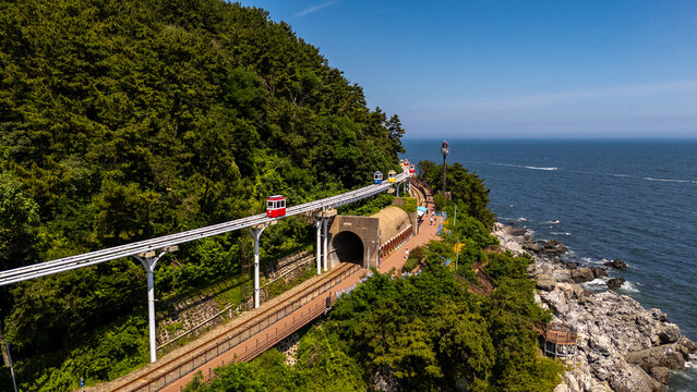 Aerial view of the Haeundae Beach Blue Line Park with colorful sky capsules traveling along an elevated track above a coastal railway tunnel in Busan, South Korea.