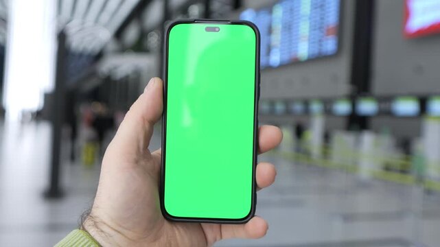 Hand holds smartphone with green screen. Airport terminal visible in background. Blurred travelers and flight info screens. Phone centered in shot. Background shows airport gates