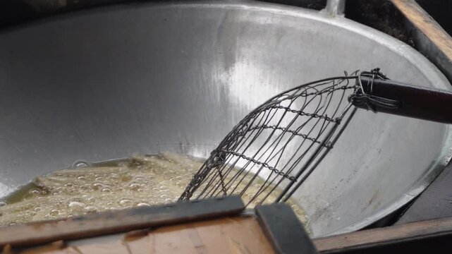 Close up of deep frying traditional snacks or dough sticks in bubbling hot cooking oil inside a large metal wok with a wire mesh strainer skimmer spoon, authentic Asian street food cooking process. So