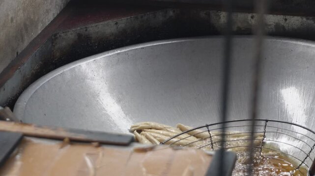 Close up of deep frying traditional snacks or dough sticks in bubbling hot cooking oil inside a large metal wok with a wire mesh strainer skimmer spoon, authentic Asian street food cooking process. So