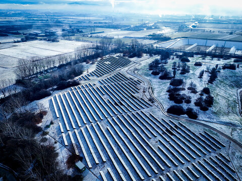Aerial view of solar park in winter with rows of photovoltaic panels covered in light snow, surrounded by frost-covered fields and trees in Druento, Piedmont, Italy.