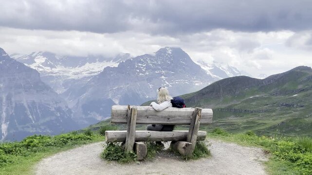 A woman traveler relaxing on a rustic wooden bench on top of a mountain, looking at the epic landscape of Eiger in Jungfrau region, Grindelwald, Switzerland