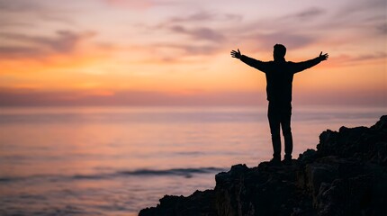 Fototapeta premium Man standing with arms outstretched on a cliff at sunset by the ocean