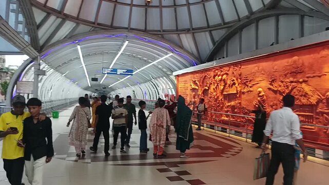 Saturday, 27 June 2026, in Kolkata, West Bengal, India. Commuters and devotees walk past a large terracotta mural inside the arched Kalighat Skywalk corridor.
