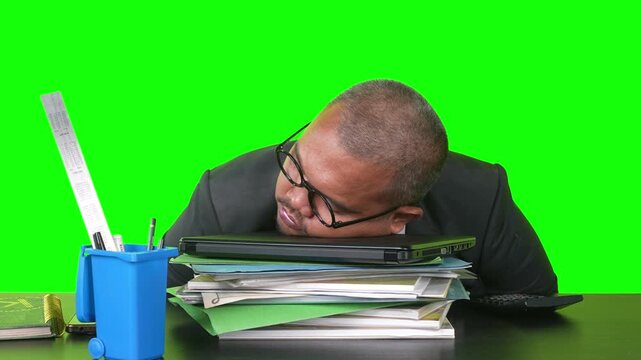 Exhausted Asian businessman sleeping on a closed laptop over a stack of paperwork at his desk against a green screen.