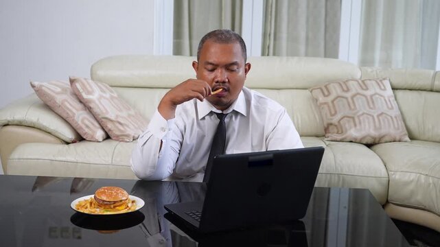Asian businessman working on a laptop while eating a fast food burger and french fries at home.