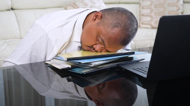 Tired Asian businessman sleeping on a stack of folders next to a laptop at home.