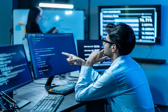 Focused software developer analyzing code on multiple computer monitors in a dark modern tech office at night. IT professional working late on cybersecurity data while colleague plans in background.