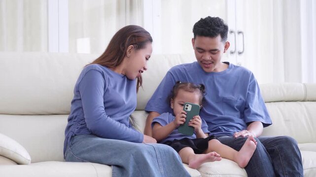 Happy Asian family with a young daughter sitting on a white sofa and looking at a smartphone together at home.