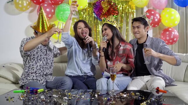 Group of joyful Asian friends singing karaoke, drinking champagne, and celebrating a birthday party together on a couch at home.