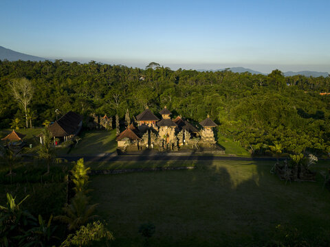Aerial view of Pura Dalem Buungan temple complex surrounded by lush tropical forest and greenery under a clear sky in Kecamatan Susut, Bali, Indonesia.