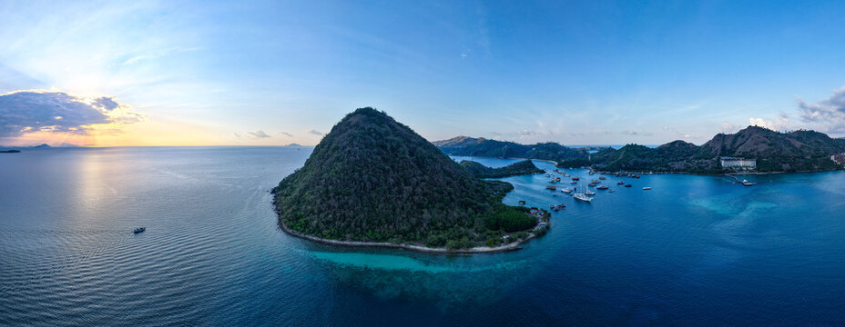 Aerial view of Pulau Kukusan with lush green hills, turquoise waters, and anchored boats at sunset, Komodo, East Nusa Tenggara, Indonesia.