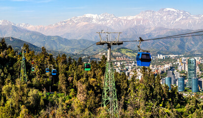 Fototapeta premium Cable cars of Teleferico Santiago glide over Santiago Metropolitan Park with the Andes mountains and city skyline in the distance.