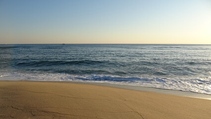 Fototapeta premium Clear Sky Sunrise over Peaceful Ocean and Sandy Beach. Beautiful morning light illuminating a quiet sandy shoreline with a distant boat on the calm horizon in Gangneung.