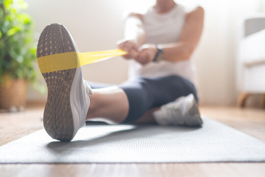 Mature woman sitting on a yoga mat on the floor at home, stretching leg muscles and strengthening arms with a yellow resistance band, focusing on personal wellness and healthy lifestyle
