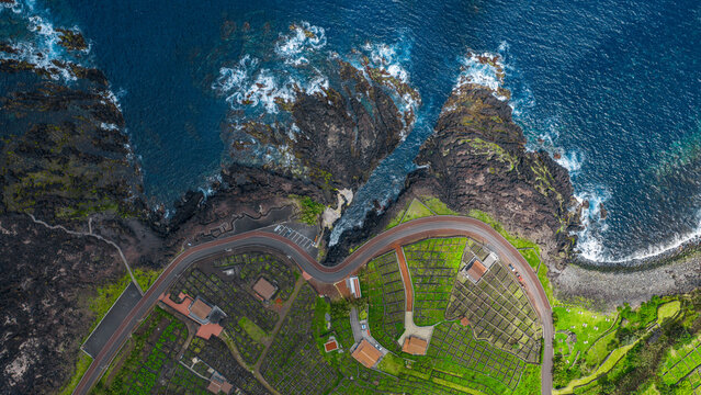 Aerial view of Piscinas Naturais Biscoitos featuring volcanic rock formations, a winding coastal road, and lush green vineyards in Biscoitos, Azores, Portugal.