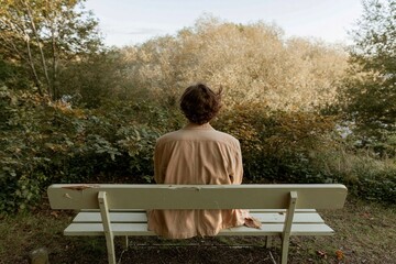 Fototapeta premium Young man seen from behind sitting on a weathered park bench gazing at autumn trees, contemplating in a peaceful nature setting for mindfulness and solitude