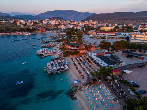 Aerial view of the coastal town of Ksamil with its turquoise waters, sandy beaches, rows of sun loungers, and surrounding hills at sunset in Ksamil, Vlore County, Albania.