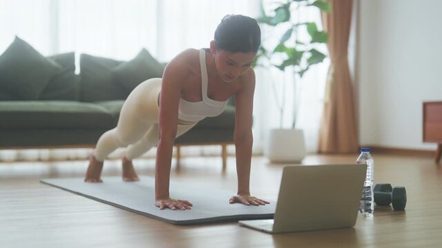 Young Asian woman in sportswear doing plank on training mat while watching online workout tutorials on laptop in the living room. Sport and recreation concept