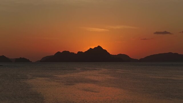 Aerial Sunset Ascent Shot at Corong Corong Beach in El Nido. Drone Vertical Climb Reveal View of Deep Orange Twilight Sky and Silhouetted Mountain Peak Over Calm Ocean.