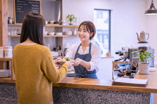 明るいお洒落なカフェのカウンターで、笑顔の女性店員がお客様に料理（ランチプレート）を両手で丁寧に提供・手渡ししている接客シーン。ビジネス、サービス、飲食店のイメージ素材