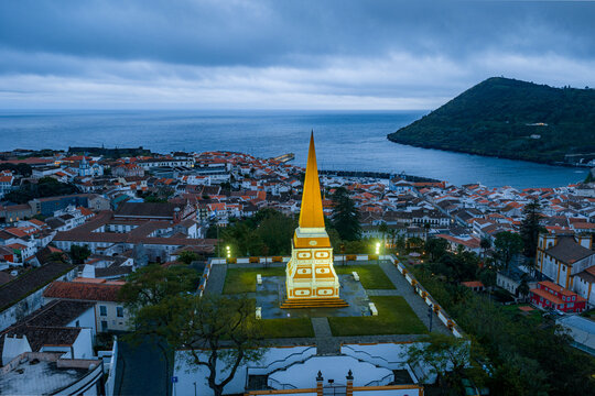 Aerial view of the illuminated Alto da Memoria obelisk overlooking the historic town with red-tiled rooftops and the Atlantic Ocean in Angra do Heroismo, Azores, Portugal.