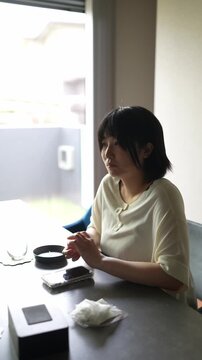 Soft light spills through a window, illuminating her quiet stillness. She gazes outward, hands folded, eyes thoughtful, calm, focused. Small bowl and phone rest on the table beside her