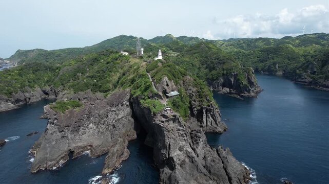 Ariel view of Japan's Izu Peninsular Cape Irozaki rugged cliffside lighthouse. Green hills surround dark stone edges. Ocean waves crash against jagged rocks below. White tower stands tall, guarding th