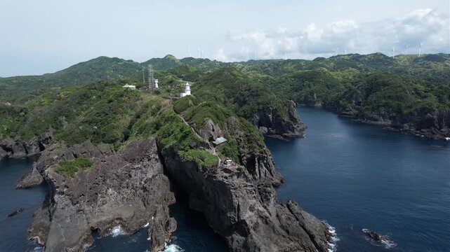 Japan's Cape Irozaki rugged cliffs rise from deep blue sea. Lone lighthouse stands firm against nature's force. Green hills frame the scene with wild, untamed beauty. Clouds drift lazily above the wil