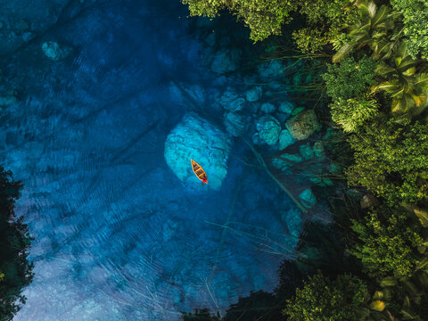 Aerial view of a small boat on the crystal clear blue waters of Danau Paisu Pok Luk Panenteng, surrounded by dense tropical forest in Bulagi Utara, Central Sulawesi, Indonesia.