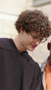 Vertical video: Turning grad in robe specs, friend in jean jacket showing phone at stone wall