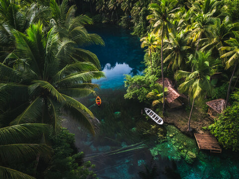 Aerial view of Danau Paisu Pok Luk Panenteng featuring crystal clear turquoise water, lush palm trees, wooden huts, and small boats in Bulagi Utara, Central Sulawesi, Indonesia.