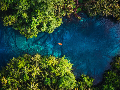 Aerial view of Danau Paisu Pok Luk Panenteng with a small orange boat on crystal clear blue water surrounded by lush tropical vegetation in Bulagi Utara, Central Sulawesi, Indonesia.