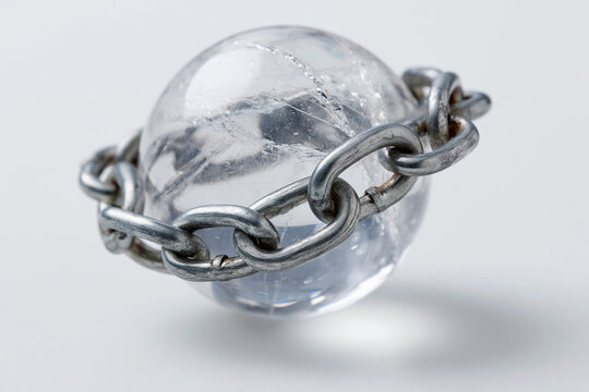 Clear crystal sphere encircled by heavy metallic chain. Chain links grip the glass with cold, industrial force. Light reflects off smooth, transparent surface. Shadow casts beneath the object